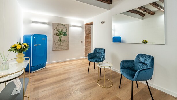Ein modernes Zimmer mit einem blauen Kühlschrank, zwei blauen Stühlen, einem kleinen runden Tisch und einem großen Spiegel an der Wand.