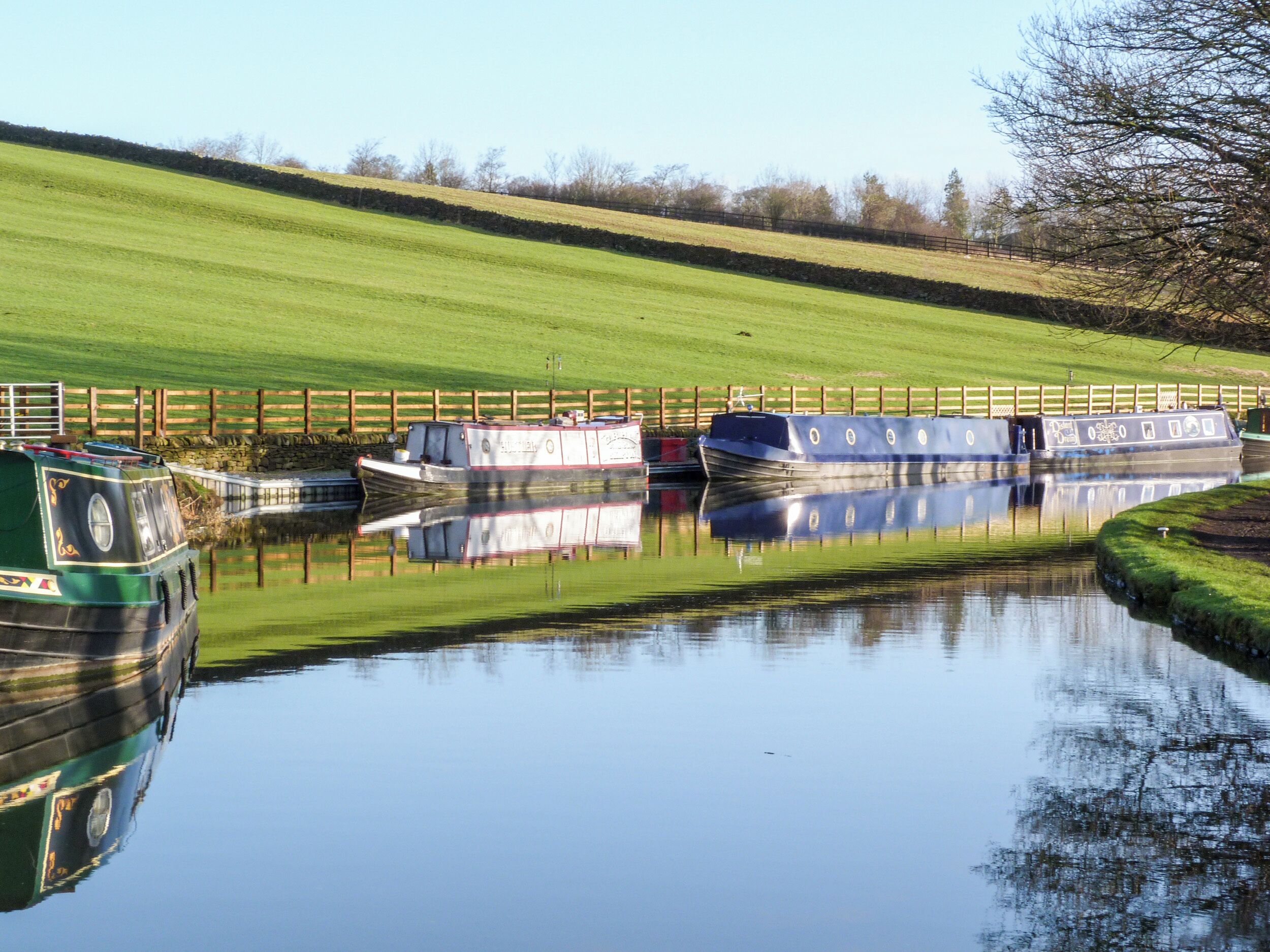 A canal with boats docked, surrounded by a grassy embankment and a wooden fence.