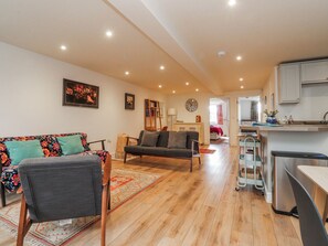 A living room with a sofa, a chair, a rug, a bookshelf, and a kitchen area with cabinets and a refrigerator. - Number 7 Canalside (Skipton)