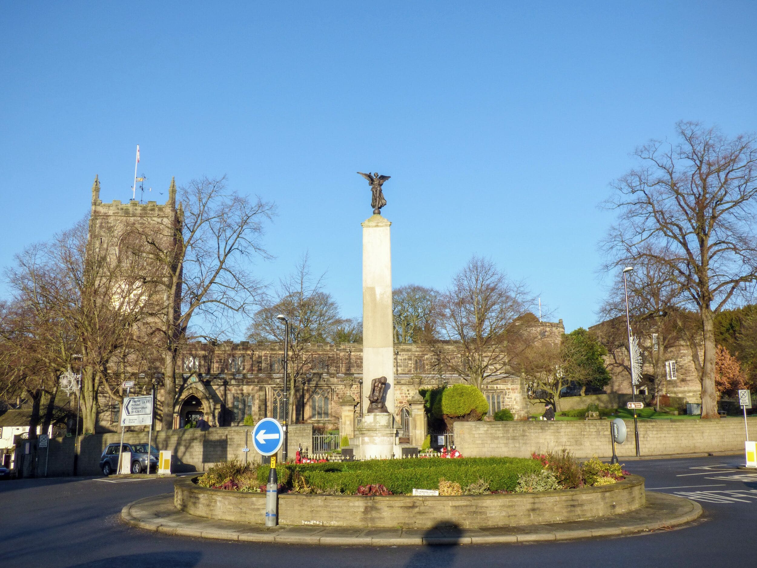 A monument with a statue, surrounded by a landscaped garden and a historic building in the background.