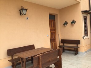 Une table et des bancs en bois se trouvent dans une pièce dont le mur est de couleur beige, avec une porte en bois et une fenêtre dotée de volets.