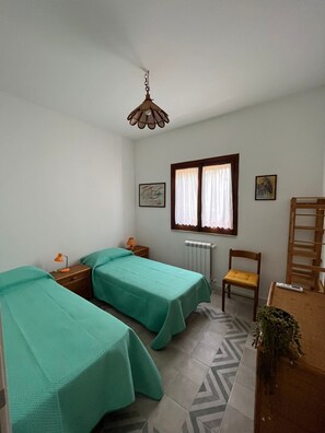 Une chambre avec deux lits simples, une chaise en bois, un bureau avec une chaise, un radiateur et une fenêtre avec des rideaux.