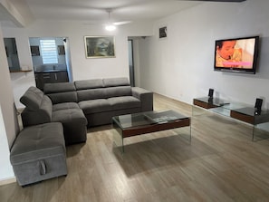 Ein modernes Wohnzimmer mit einem Ecksofa, einem Glastisch, einem Flachbildfernseher und einer Küche im Hintergrund.