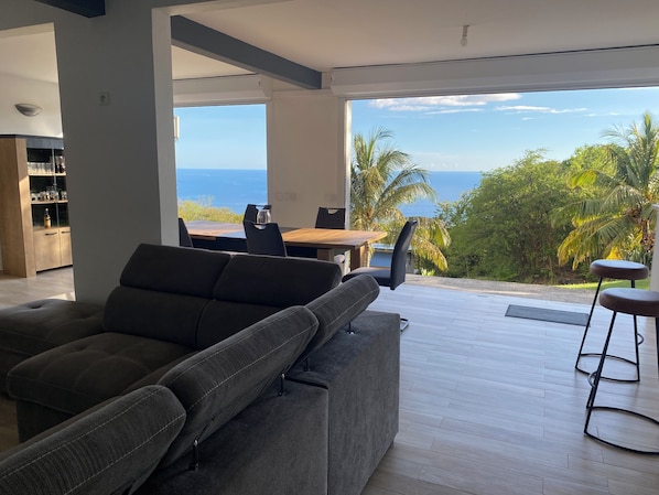 Ein modernes Wohnzimmer mit einer großen Couch, einem Esstisch und Stühlen. Durch große Fenster ist der Blick auf den Ozean und Palmen zu sehen.