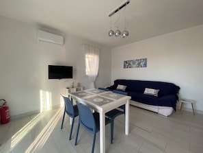 A modern living room with a white sofa, a dining table with chairs, a flat-screen TV mounted on the wall, and a ceiling light fixture. - Fabulous 2-bedroom apartment in La Ciaccia (La Ciaccia)