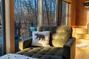 A room with a large window, a cushioned armchair, a pillow with a bear design, and a wooden interior.