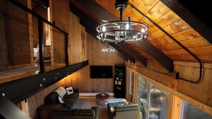 A loft-style room with a high ceiling, wooden beams, a modern chandelier, a sofa, a coffee table, and a view through large windows.