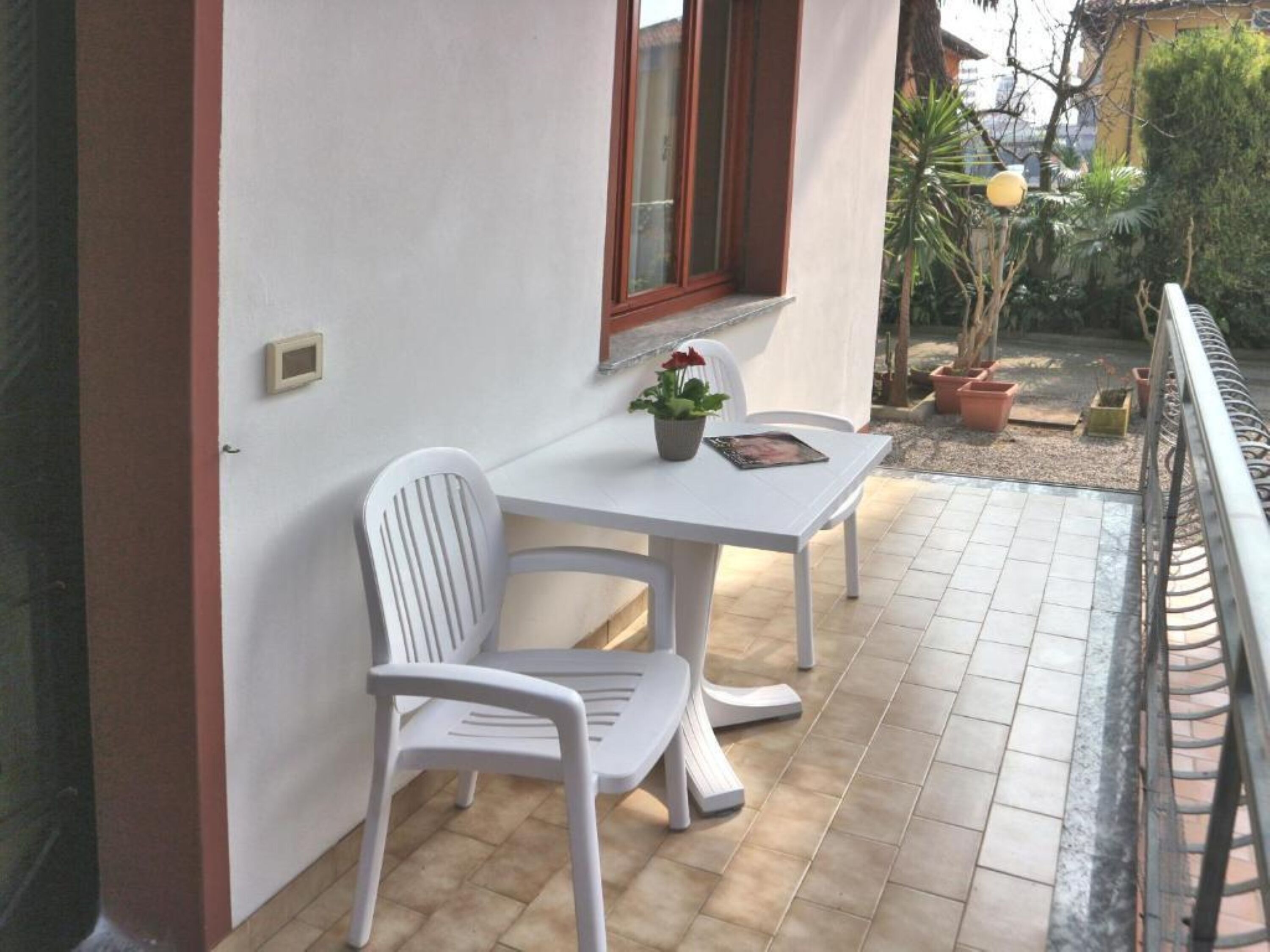 A small outdoor patio with a white table and chairs, a potted plant, and a window with a red frame.