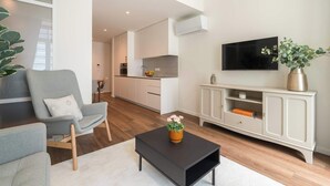 Ein modernes Wohnzimmer mit einem grauen Sessel, einem schwarzen Couchtisch mit einer Vase, einem Fernseher an der Wand und einer Küche im Hintergrund.