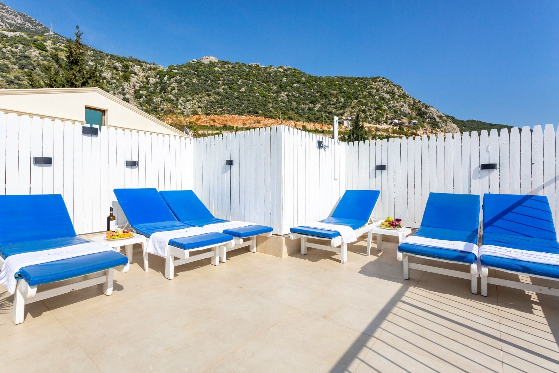 A sunlit outdoor seating area with white and blue lounge chairs, a tiled floor, and a white fence.