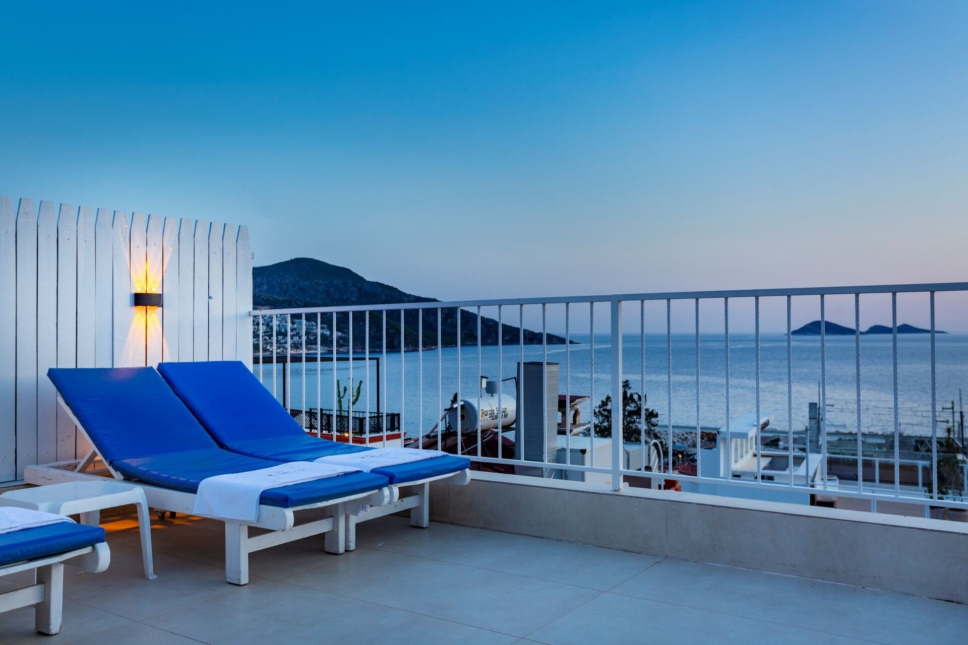 Un balcón con dos sillones azules, barandilla blanca y vista al mar y montañas distantes.
