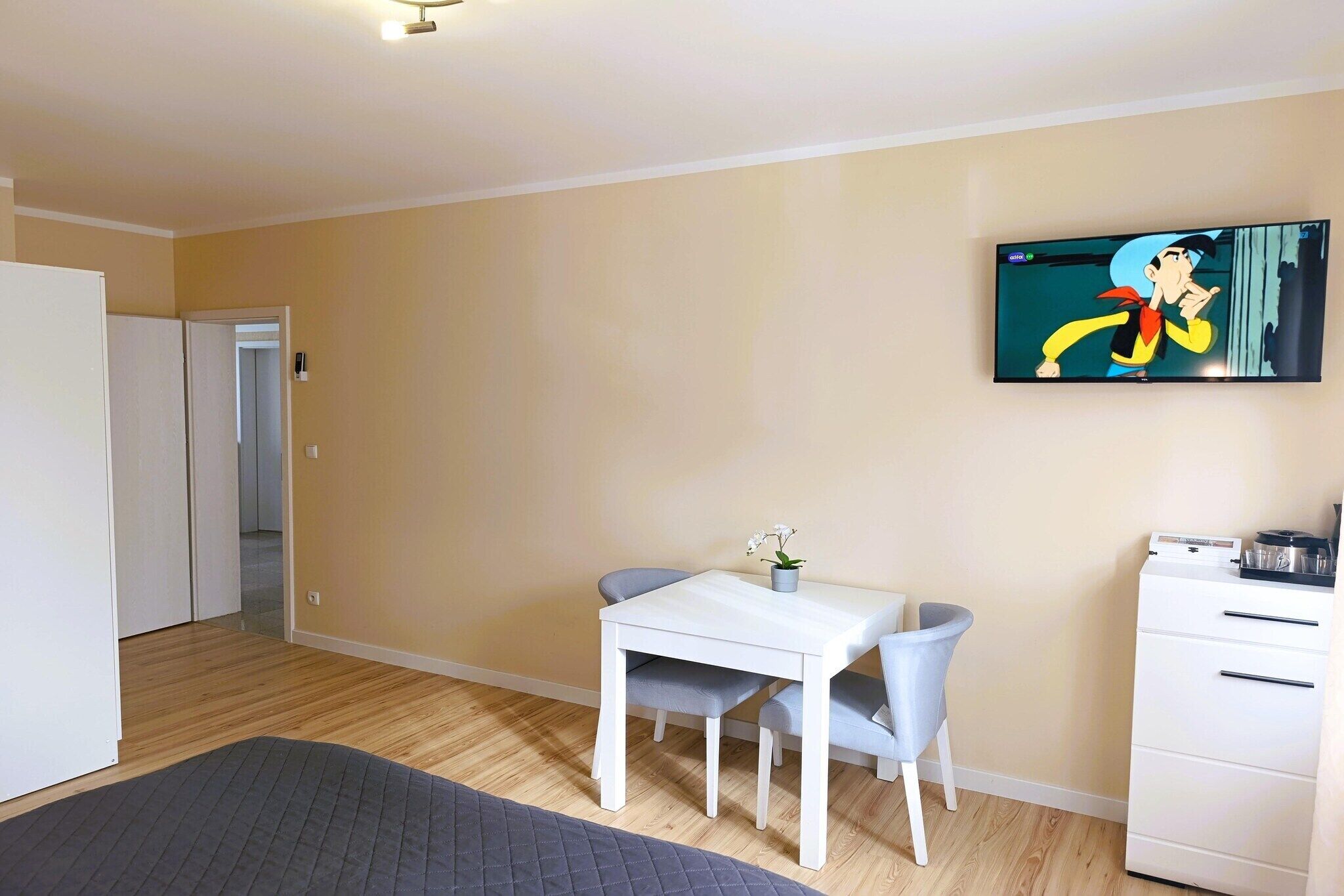 A room with a white desk, two chairs, a television on the wall, a wardrobe, and a bed with a gray bedspread.
