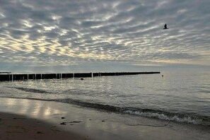 Ein Strand mit einem Steg, der sich ins Wasser erstreckt, sanfte Wellen am Ufer und ein bewölkter Himmel.