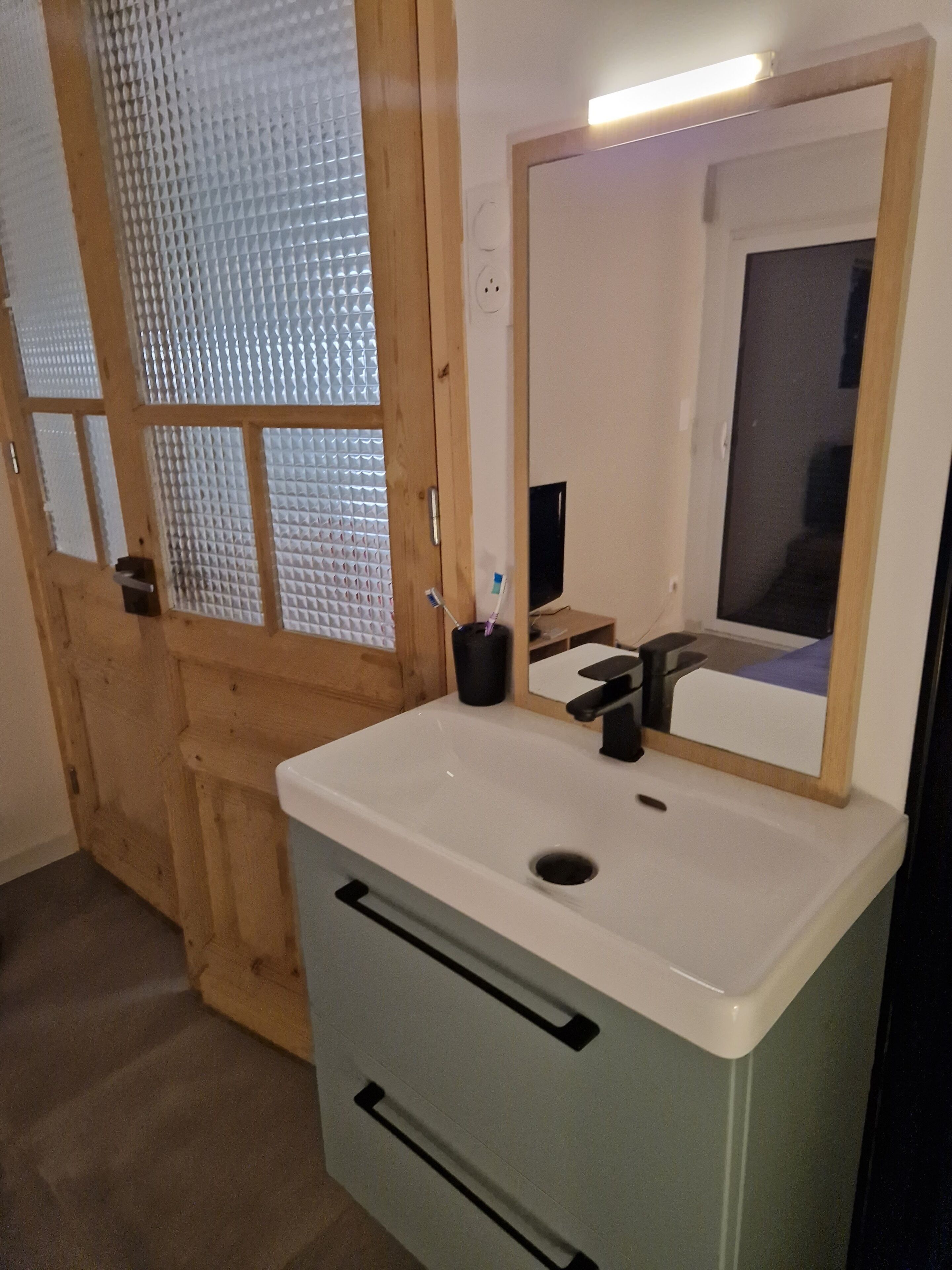 Een badkamer met een wit wastafelmeubel, een spiegel en een houten deur met geëtste glaspanelen.