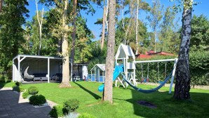 Un parque infantil con tobogán y columpios, rodeado de árboles y una zona de estar cubierta.