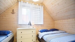 Una habitación pequeña y acogedora con techo inclinado, un aparador de madera con cajones, una cama con ropa de cama a rayas azules y blancas, y una ventana con cortinas blancas.