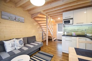 A cozy cabin interior with a wooden staircase, a sofa with nautical-themed pillows, a kitchen area with a microwave, and a wooden floor.