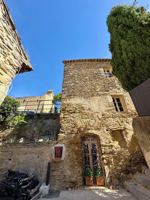 Un bâtiment en pierre doté d’une petite fenêtre et d’une porte, entouré de verdure et d’autres bâtiments.