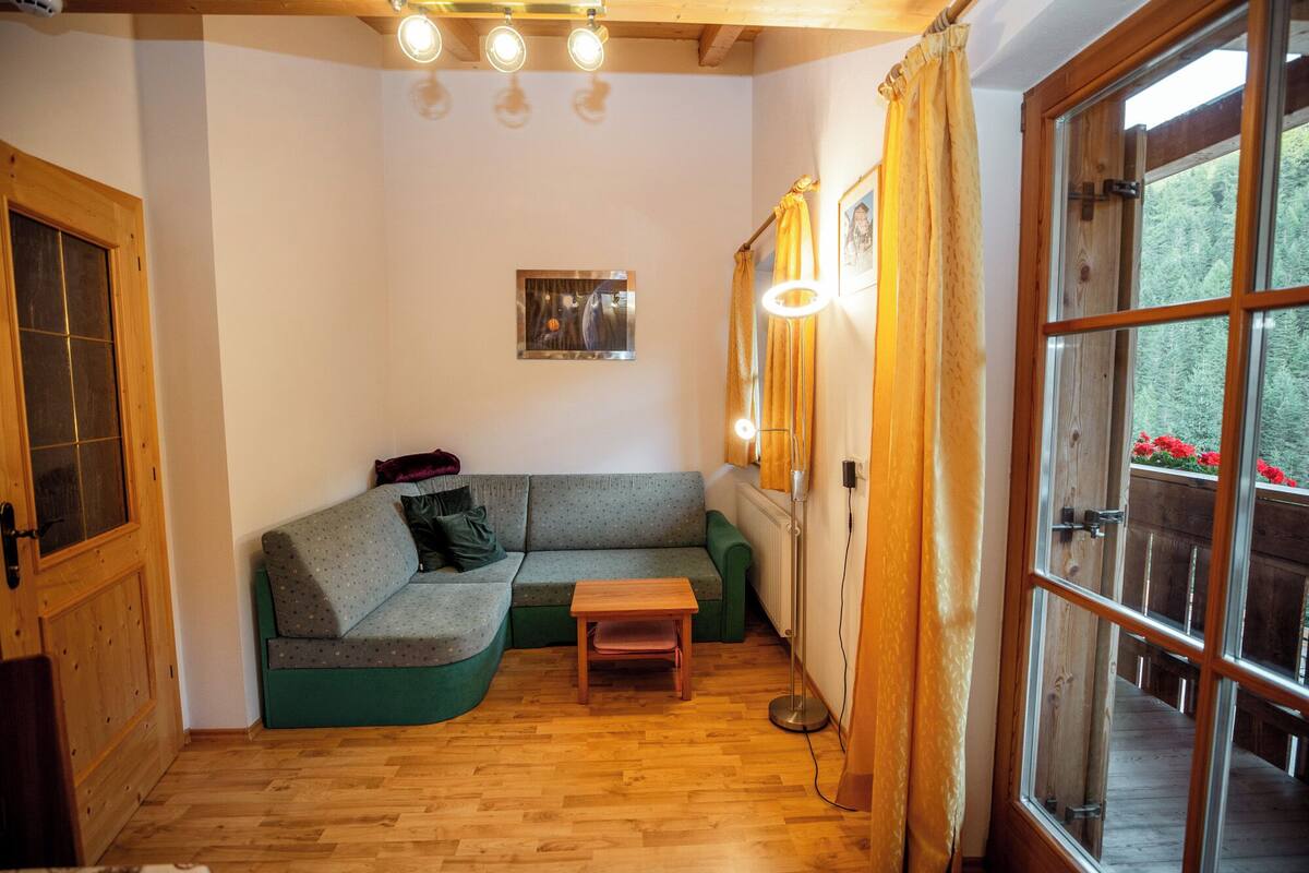 Ein gemütliches Zimmer mit Holzboden, einem grünen Sofa, einem kleinen Tisch und einem Fenster mit Vorhängen.