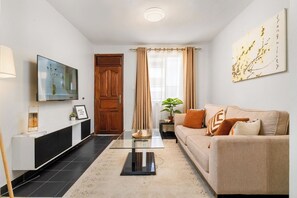 Ein Wohnzimmer mit einer Couch, einem Couchtisch, einem Fernseher, einer Holztür und einem Fenster mit Vorhängen.