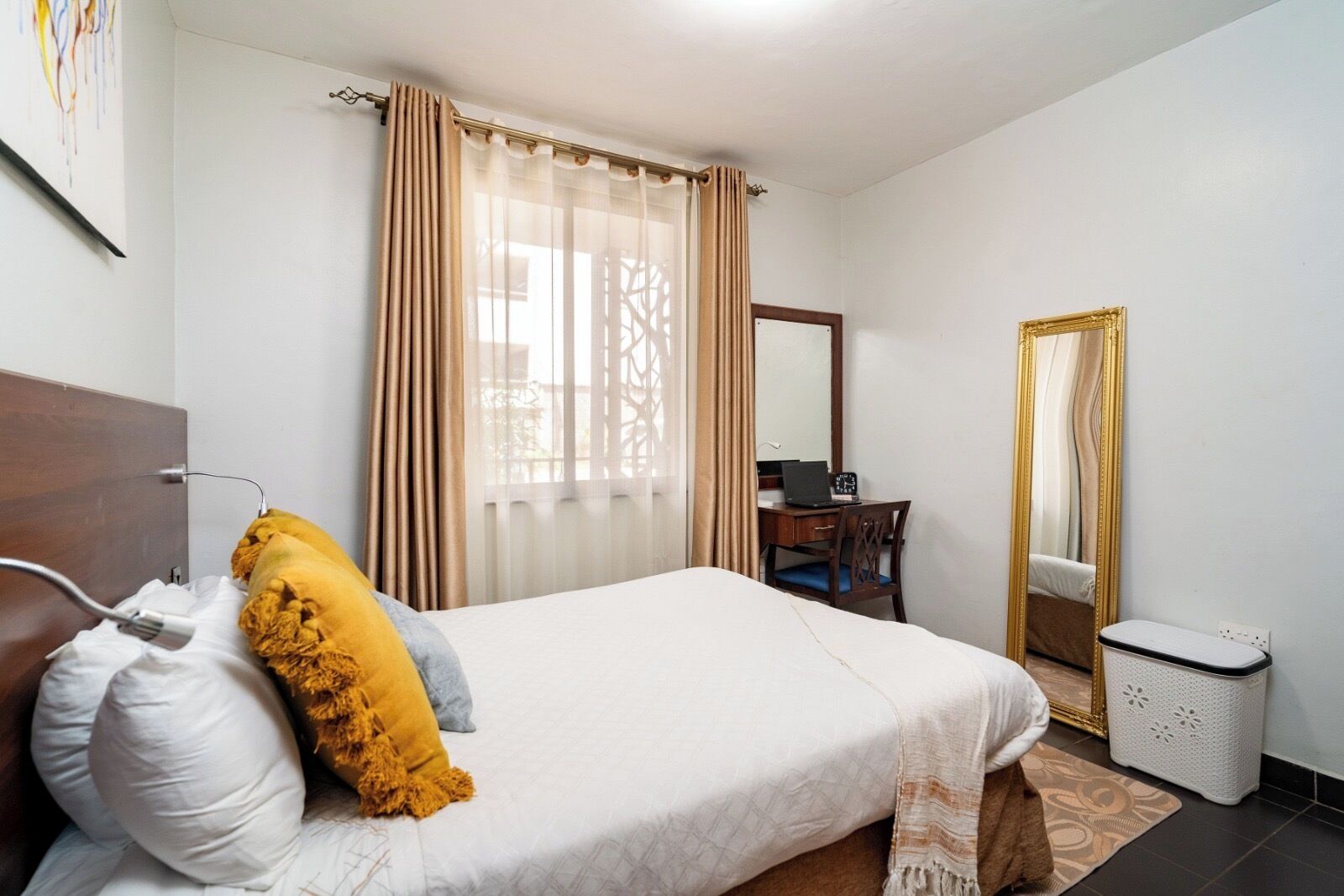 A hotel room with a bed, a desk with a laptop, a mirror, and a trash bin.