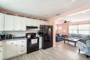 A modern kitchen with white cabinets, a black refrigerator, and a stove. There is a living area with a sofa and a coffee table. - A piece of sunshine (Bradenton)