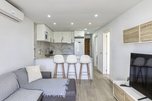 Una sala de estar moderna con un sofá gris, un televisor y una cocina con armarios blancos y un salpicadero de mármol.
