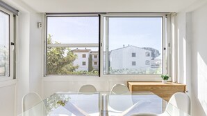 Un'area pranzo moderna con un tavolo in vetro e sedie bianche, ampie finestre con vista su edifici e alberi, e un mobiletto in legno con una pianta.