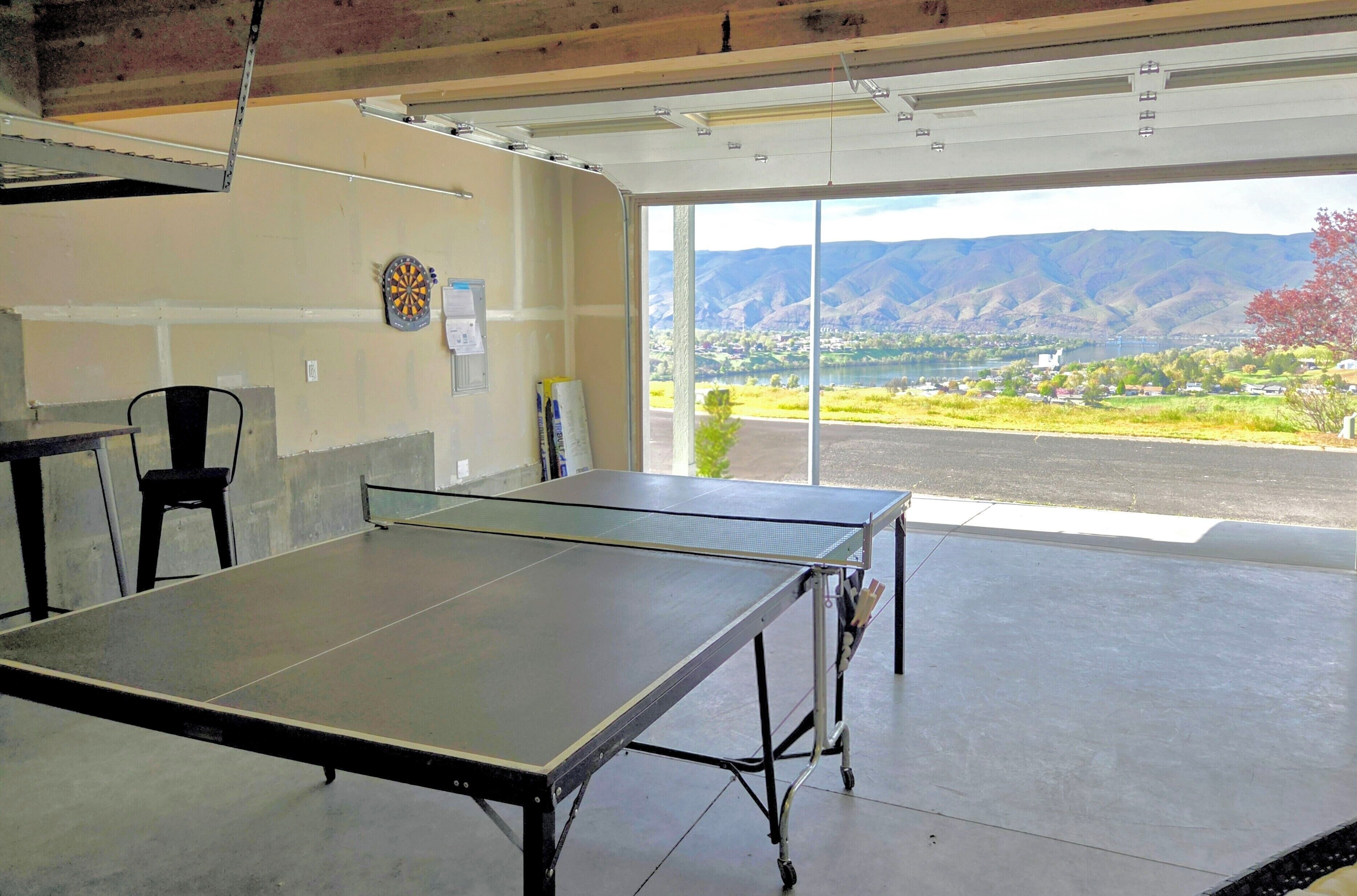 Una mesa de ping pong en una habitación con un ventanal grande, un dardo y una silla.