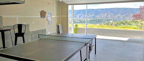 Una mesa de ping pong en una habitación con un ventanal grande, un dardo y una silla.