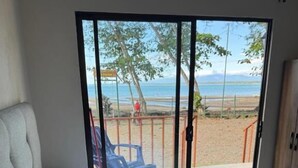 Een bed met witte lakens en groene kussens, uitzicht op het strand, en een schuifdeur naar een buitenruimte met een stoel.