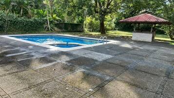 A swimming pool with a tiled deck, surrounded by lush greenery and a gazebo.