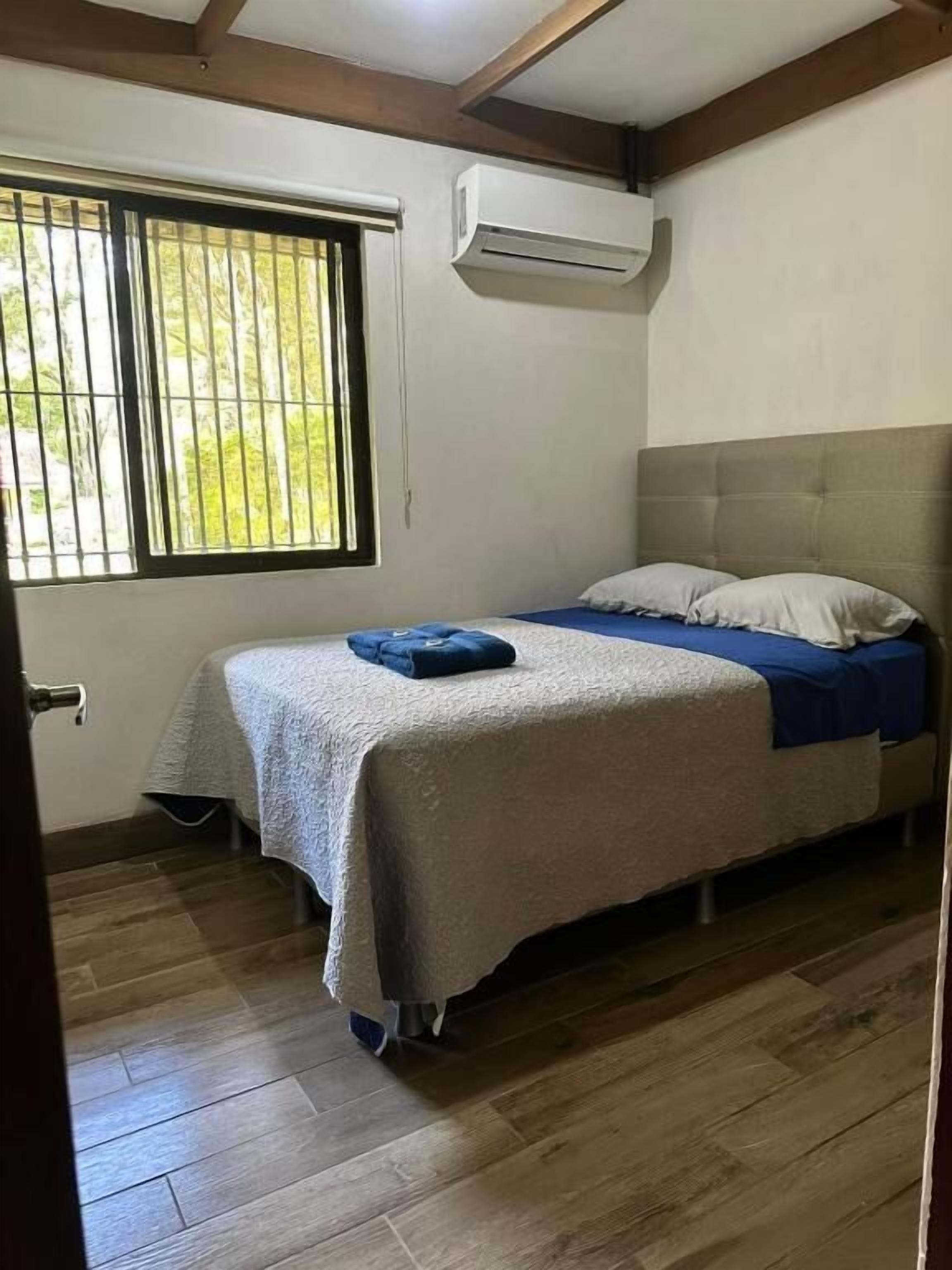 Un dormitorio con cama, ventana con rejas, equipo de aire acondicionado y suelo de madera.