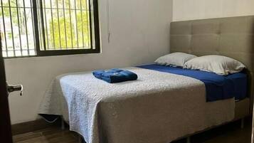 Un dormitorio con cama, ventana con rejas, equipo de aire acondicionado y suelo de madera.