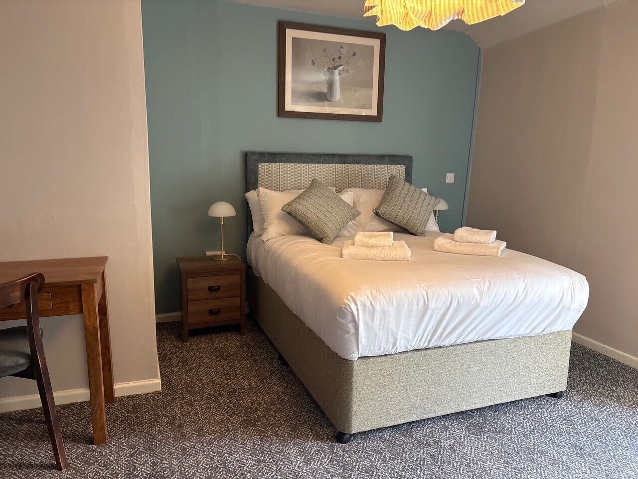 A neatly made bed with pillows and a bedside table with a lamp, in a room with a patterned carpet and a framed picture on the wall.