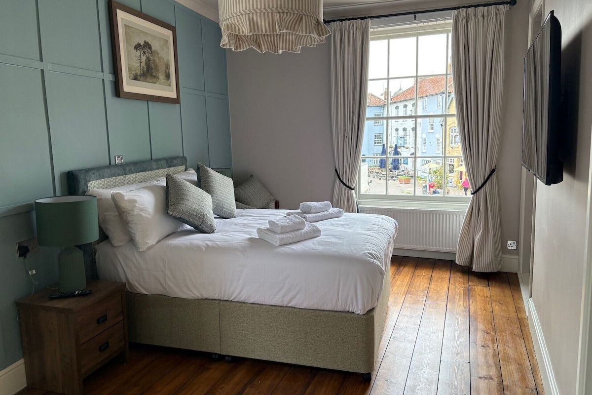Ein Schlafzimmer mit einem Bett, einem Nachttisch, einer Lampe, einem Fenster mit Vorhängen und einem Bild an der Wand.