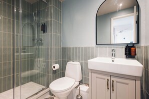 A bathroom with a glass-enclosed shower, a toilet, a sink with a mirror above it, and a roll of toilet paper on the wall.