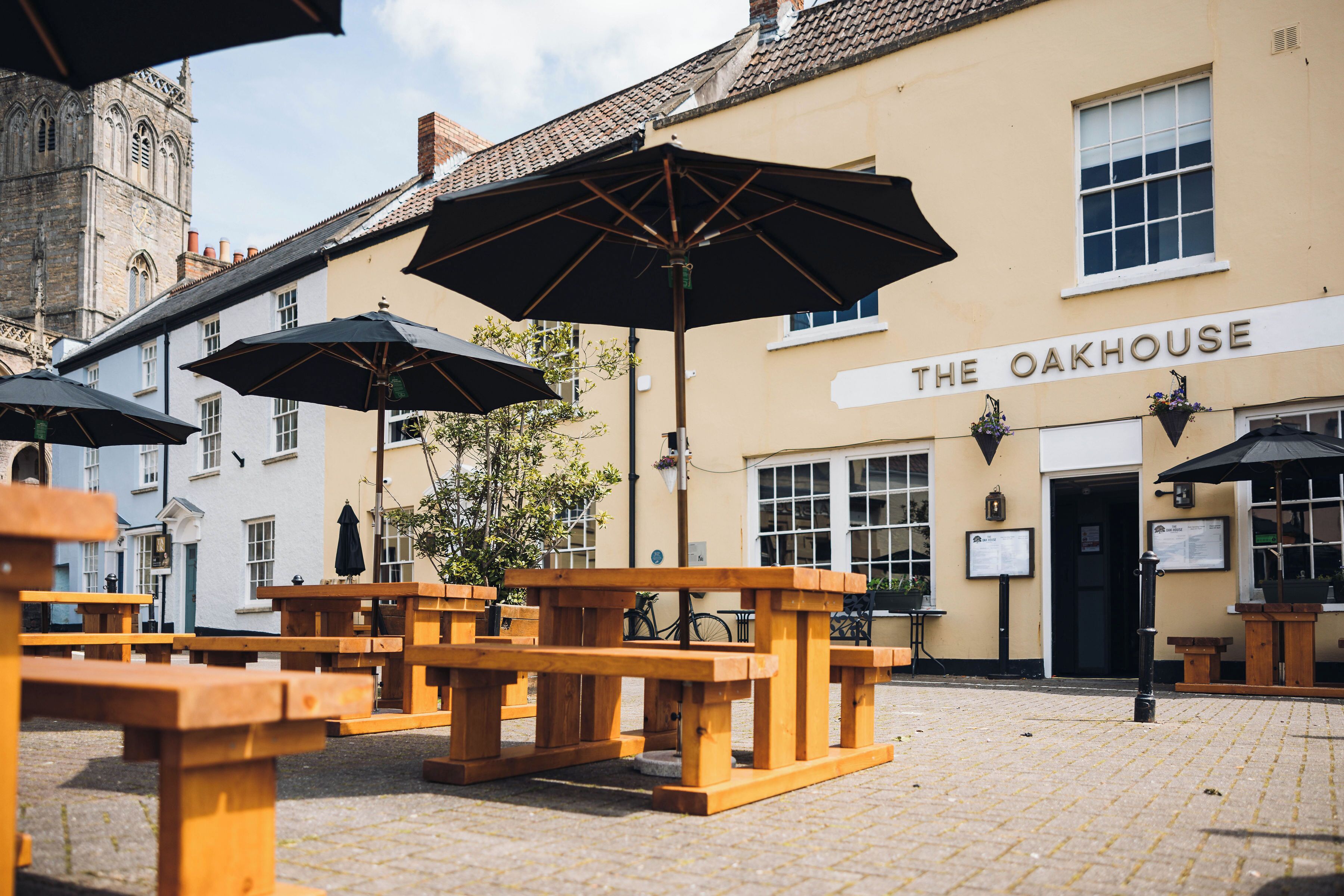 Un espace extérieur aménagé avec des bancs en bois et des parasols noirs, situé devant le restaurant The Oakhouse.
