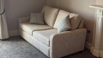 Un sofá color beige con dos almohadas y una alfombra estampada en una habitación con pared blanca y un mueble pequeño blanco.