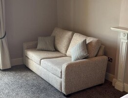 Un sofá color beige con dos almohadas y una alfombra estampada en una habitación con pared blanca y un mueble pequeño blanco.