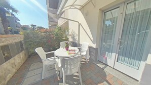 A small outdoor patio with a round table and white plastic chairs, a potted plant, and a sliding glass door. - Practical Family Base in a Lovely Area (Lignano Sabbiadoro)