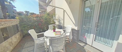 A small outdoor patio with a round table and white plastic chairs, a potted plant, and a sliding glass door.
