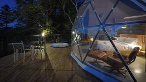 A transparent dome structure with a bed, a lounge chair, and a dining table on a wooden deck. - Bel Air Tremblant DÔME CLASSIQUE: 1 LIT KING, JACUZZI ET SAUNA-450PI (La Conception)