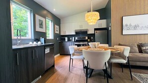 A modern kitchen with dark cabinets, a dining area with a round table and chairs, and a large window. - Bel Air Tremblant L'ETOILE : 2 CHAMBRES, JACUZZI PRIVÉ (La Conception)
