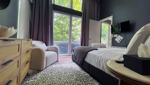 A bedroom with a bed, a dresser, a chair, a bedside table, a TV, and large windows with a view of trees. - Bel Air Tremblant L'ETOILE : 2 CHAMBRES, JACUZZI PRIVÉ (La Conception)