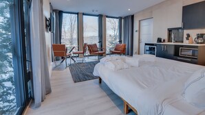 A modern hotel room with a large bed, a seating area with chairs, a kitchenette, and large windows offering a view of trees and mountains.