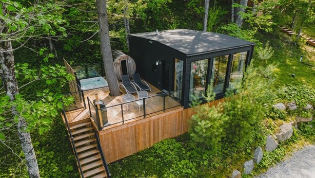 A modern cabin with a wooden deck, a glass-enclosed room, and a hot tub.