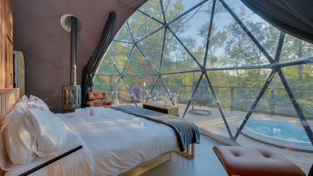 A bedroom with a bed, a bedside table, a heater, and a view through a large window. - Bel Air Tremblant DÔME MEZZANINE : 1 KING, 2 SIMPLE, JACUZZI, SAUNA-450PI (La Conception)