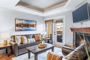 A living room with a sofa, coffee table, fireplace, television, and a painting on the wall. - Lovely 2BR Rockies Getaway w Pool + Hot Tub Access (Canmore)