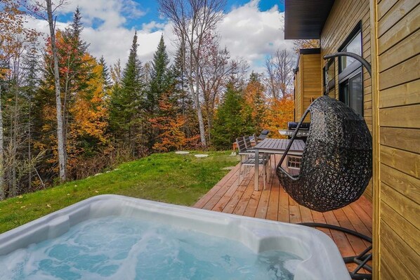 A hot tub on a wooden deck with a table and chairs, surrounded by trees with autumn foliage.